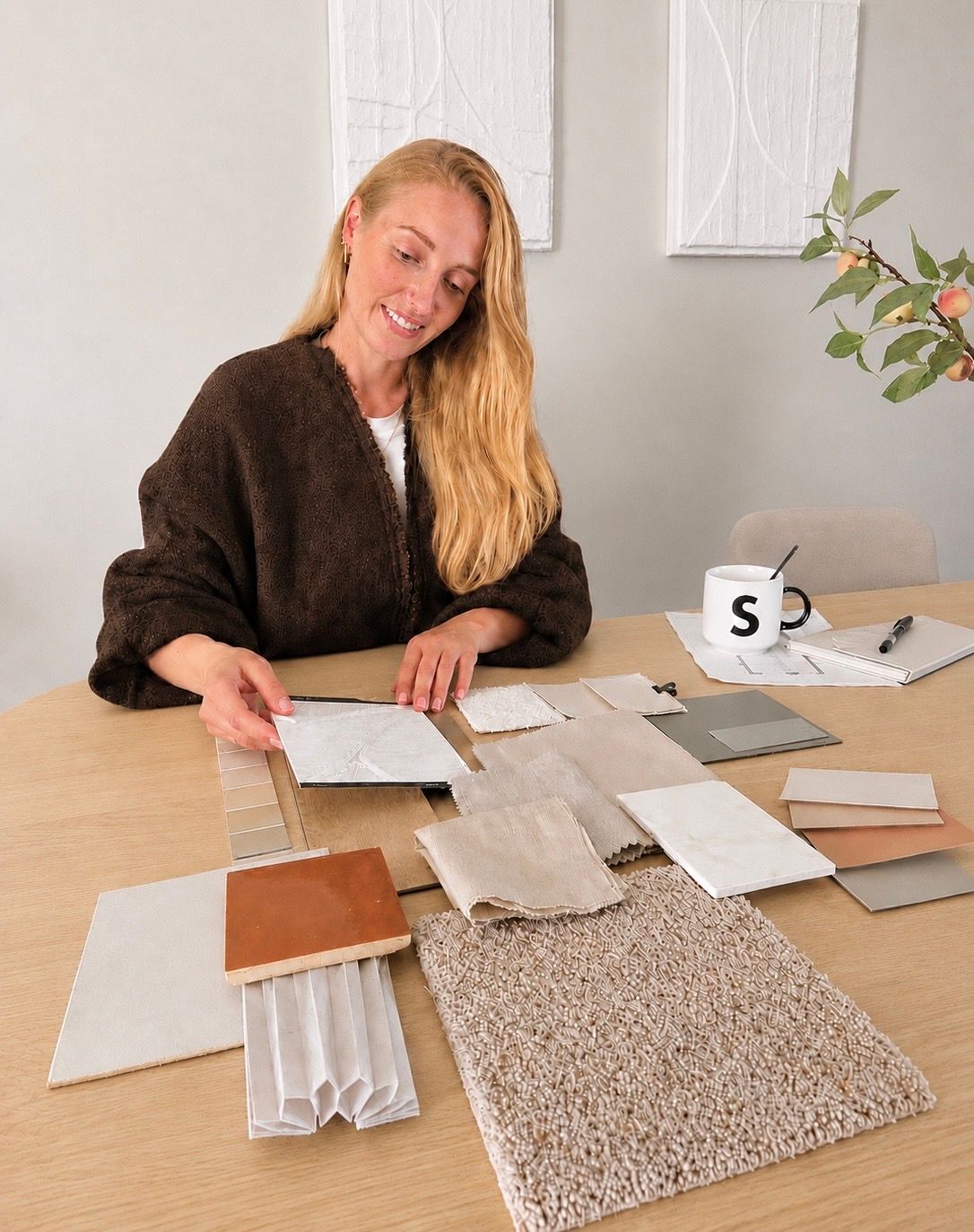 Suzanne zit aan haar werktafel en heeft voor haar een levend moodboard liggen met materiaal en kleurstalen. Denk aan diverse stofstalen en passende verfstalen. Suzanne houdt een behanghaal staal vast.